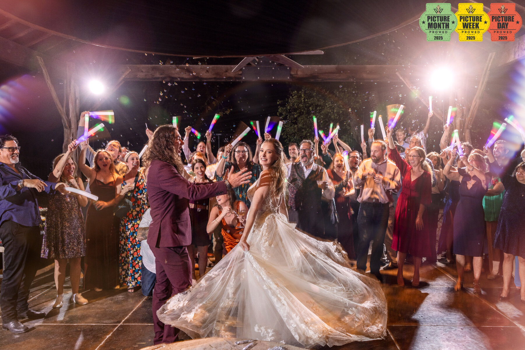Bride twirls on dance floor surrounded by cheering guests with glow sticks during nighttime wedding celebration.