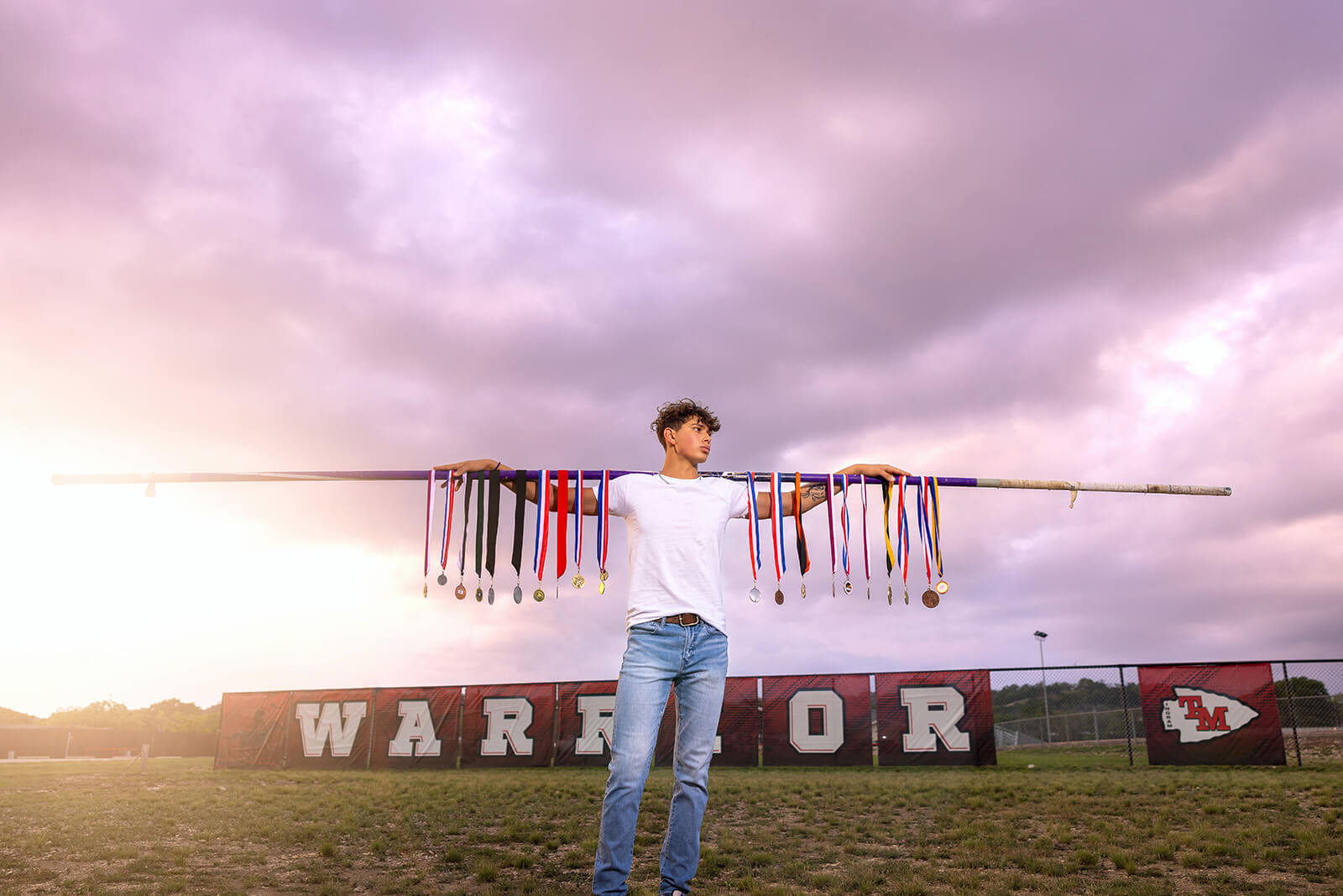 High school senior athlete holding medal-covered pole vault bar in front of 'Warrior' banner on track field in Ingram, Texas during sunset